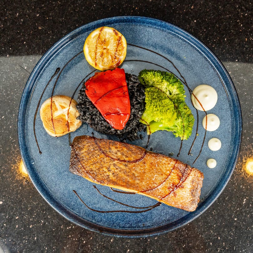 Grilled salmon with black rice, broccoli, roasted red pepper, mushroom, and lemon on a blue plate.
