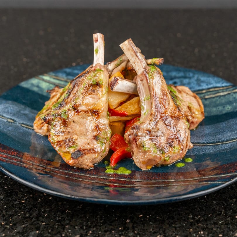 Grilled lamb chops with green sauce, roasted potatoes, and red peppers on a blue plate.